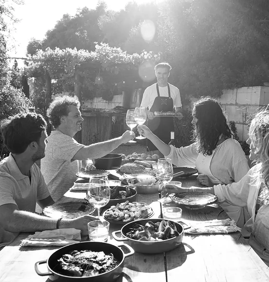 Een groep mensen geniet van een gezellige buitentafel vol met eten en COMBEKK-pannen terwijl ze toosten in de zon.