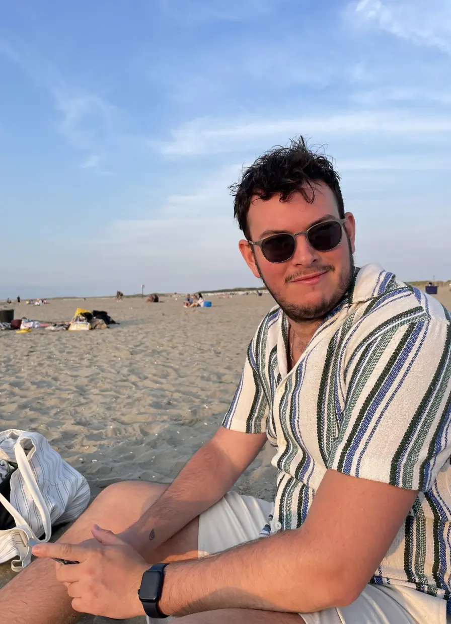 Joris zit ontspannen op het strand met een gestreept shirt en zonnebril, terwijl de zon ondergaat.