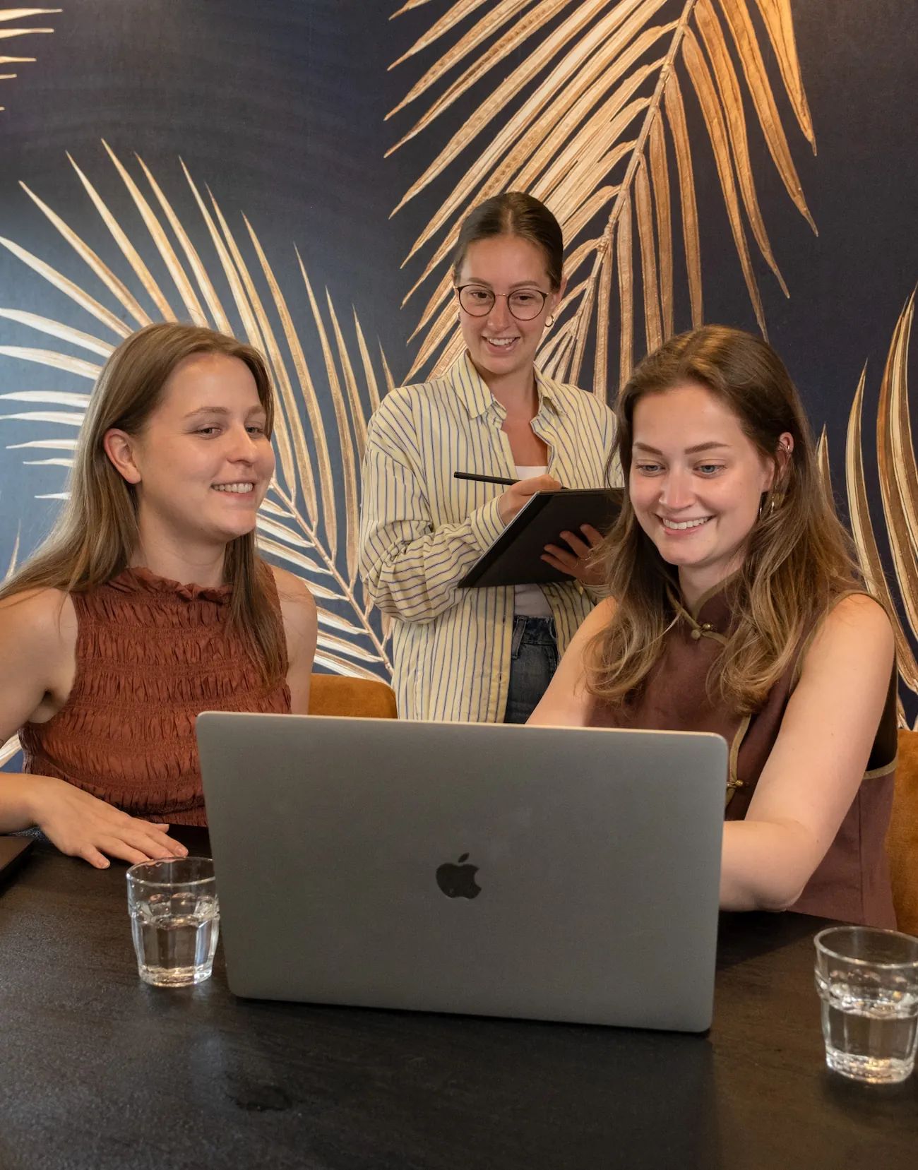 Drie jonge vrouwen overleggen lachend over marketing achter een laptop in een modern kantoor.