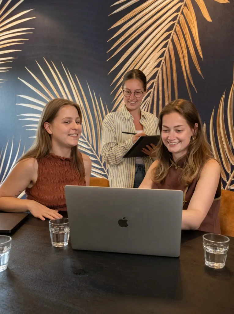 Benthe, Isabelle en Lotte werken samen met een laptop en notitieblok aan een tafel in een modern kantoor.