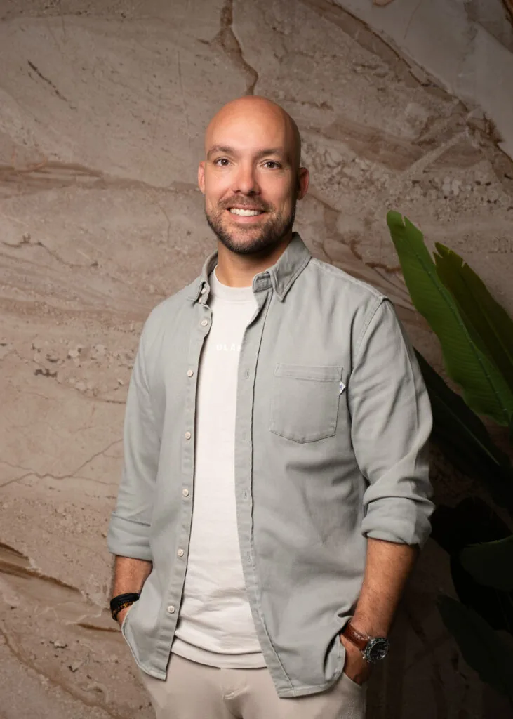 Man met een kaal hoofd en baard glimlacht naar de camera voor een marmerachtige muur met een groene plant.