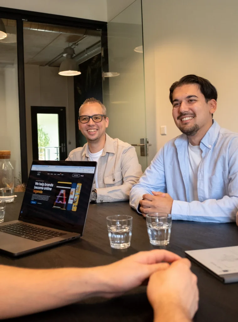 Twee glimlachende mannen in een vergaderruimte zitten aan tafel tegenover een collega met een open laptop.